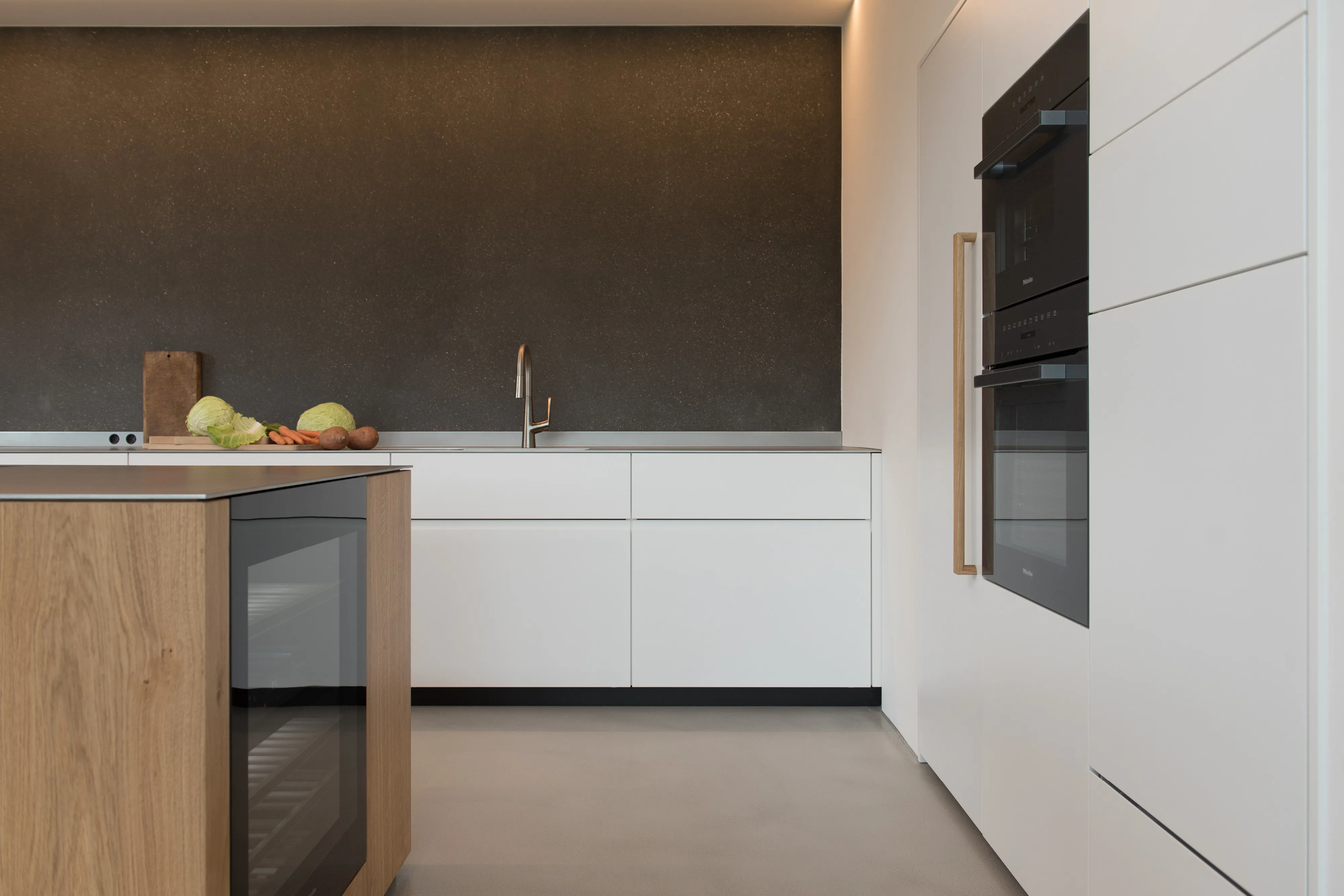 Nahaufnahme der Küchenzeile mit weißer Front, integrierten Backöfen, Holzkühlschrankgriff und seitlichem Weinkühlschrank