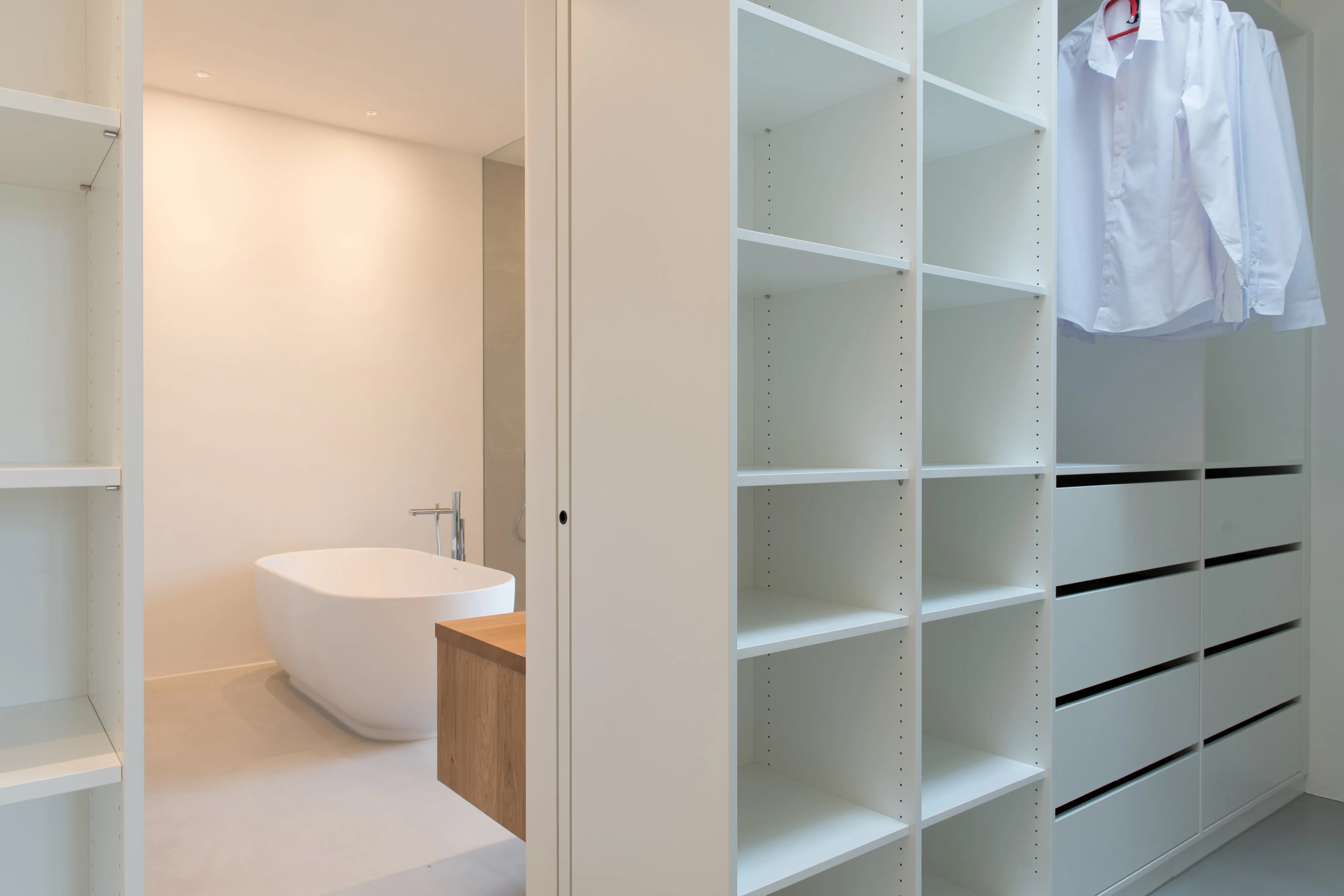 Blick vom begehbaren Kleiderschrank mit weißen Einbauregalen in das angrenzende Badezimmer mit freistehender Badewanne