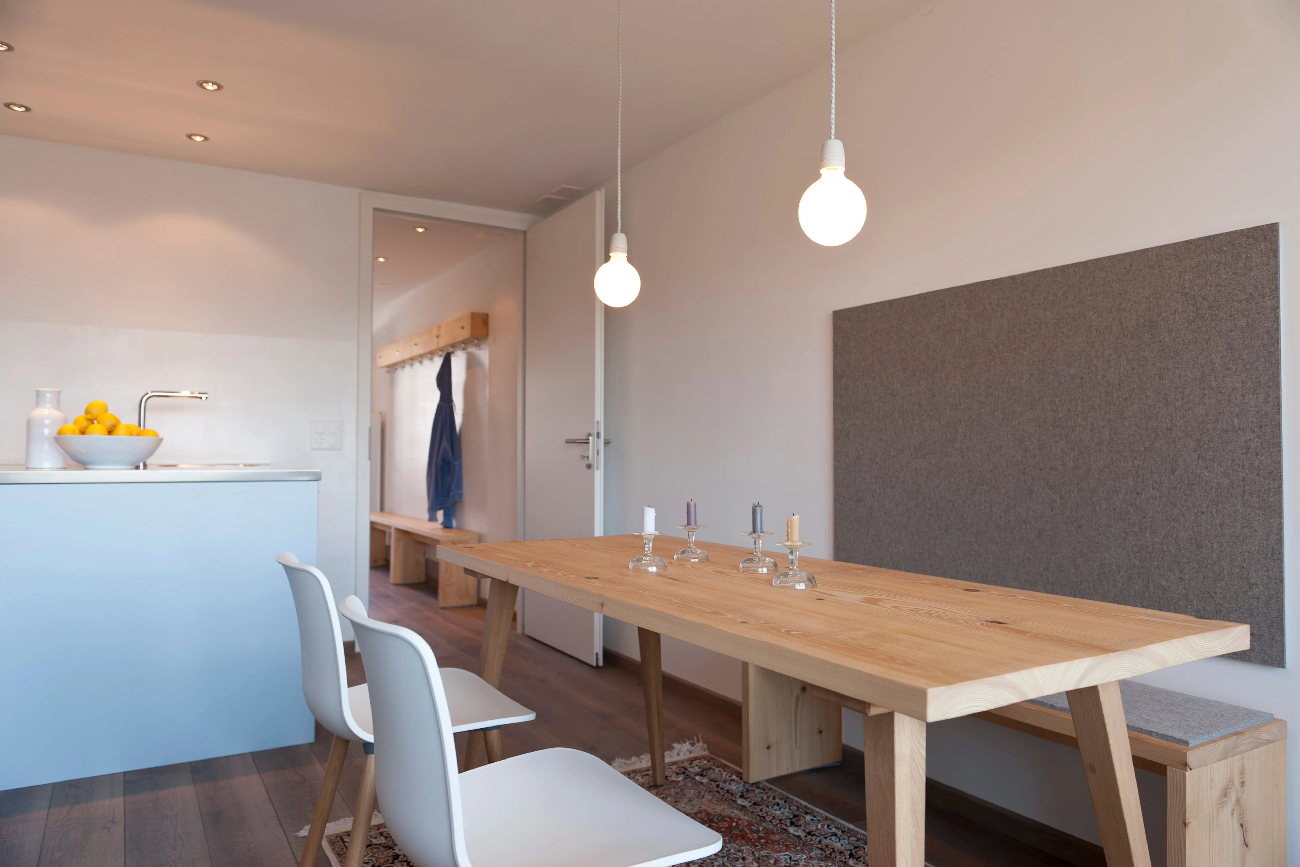 Helles Esszimmer mit Holztisch, Sitzbank, Filzwandpaneel und Pendelleuchten neben einer hellblauen Kücheninsel