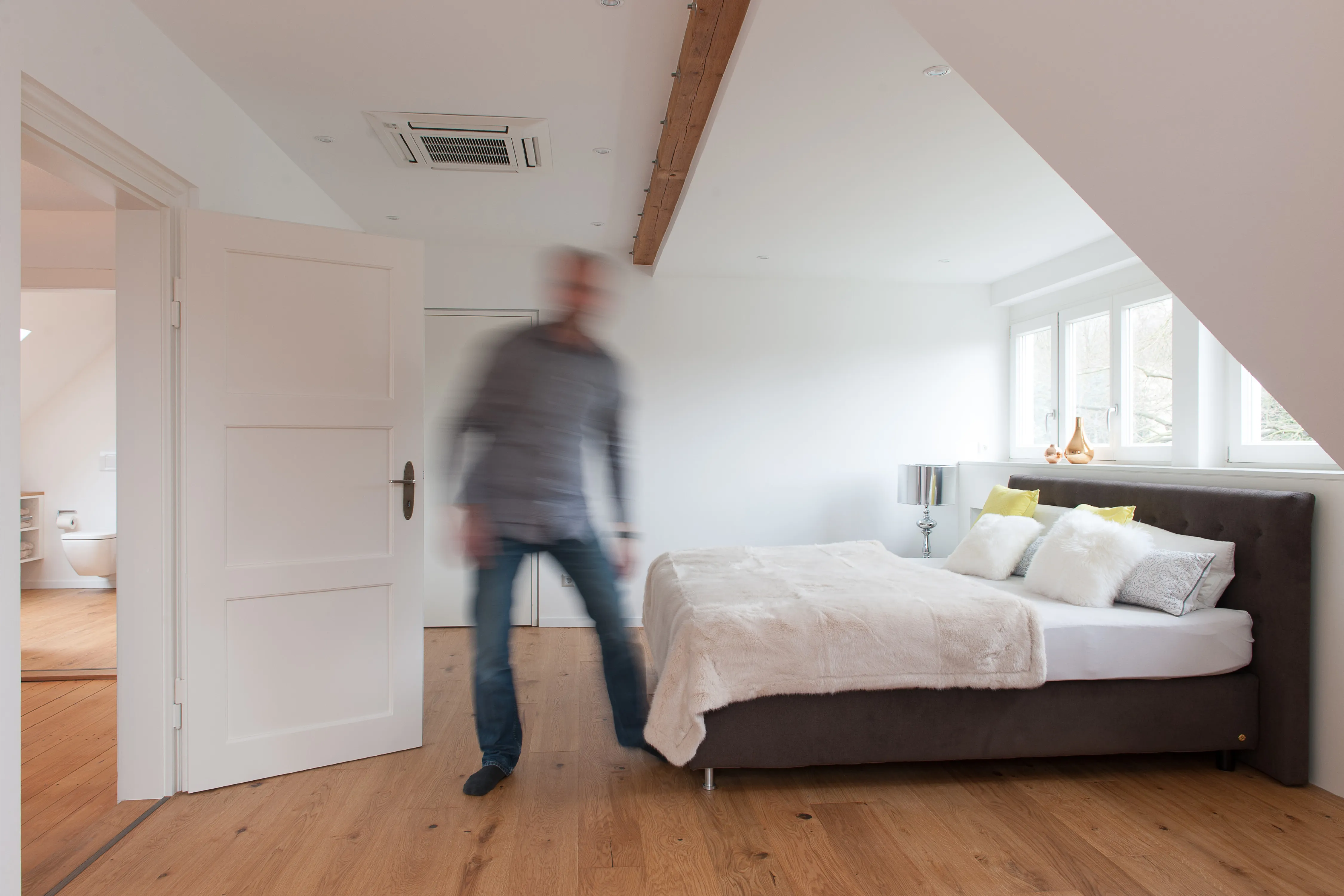 Helles Schlafzimmer unter dem Dach mit sichtbaren Holzbalken und einer hindurchgehenden Person mit Bewegungsunschärfe im Vordergrund.