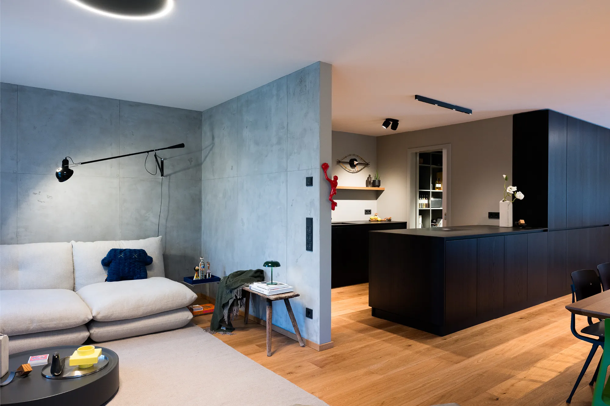 Moderner Wohnbereich mit hellem Sofa, Betonoptik-Trennwand und Übergang zur dunklen Einbauküche.