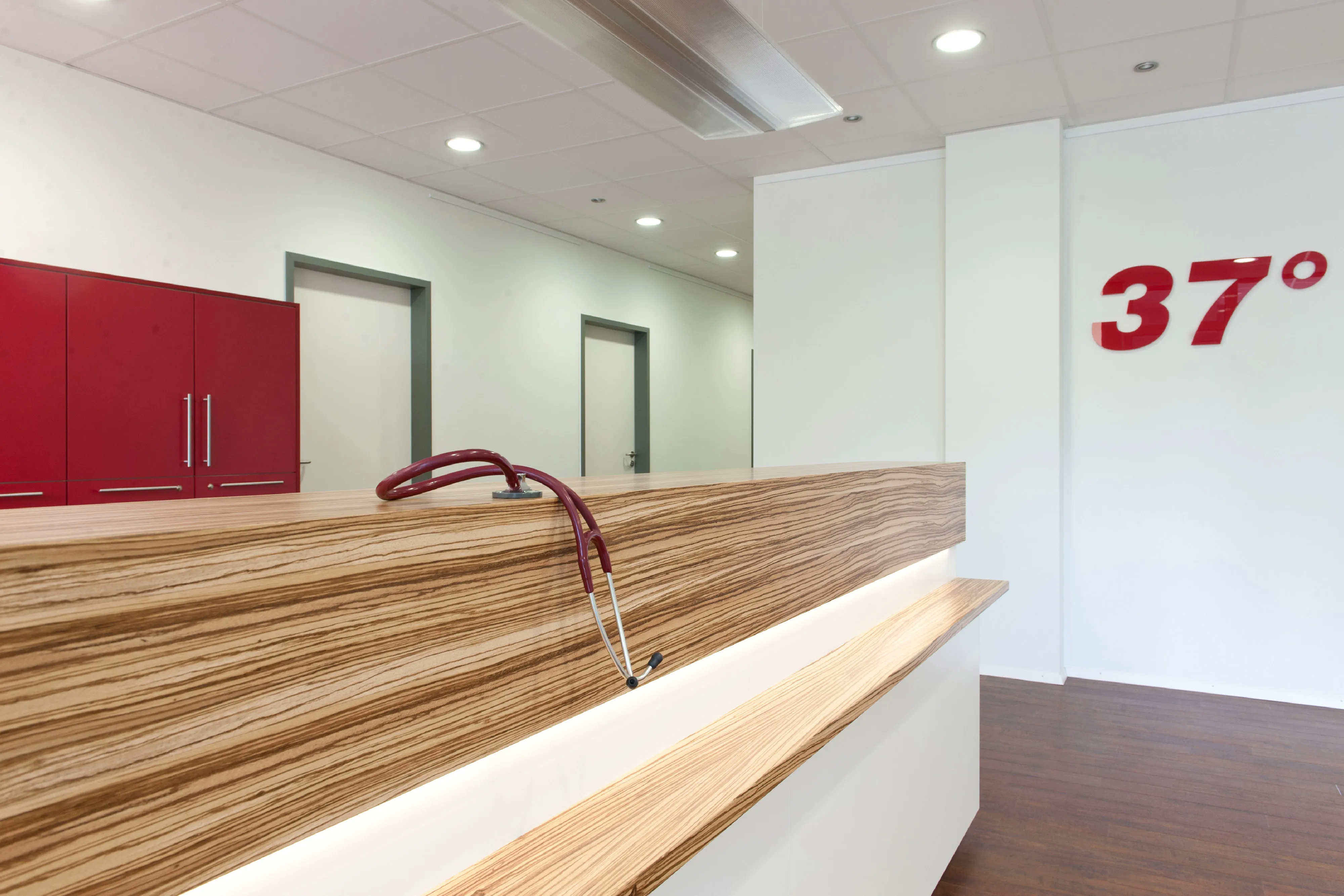 Empfangstheke in Holzoptik mit Zebranomuster und integrierter Beleuchtung, darauf liegt ein Stethoskop vor dem Praxisschriftzug „37°“.