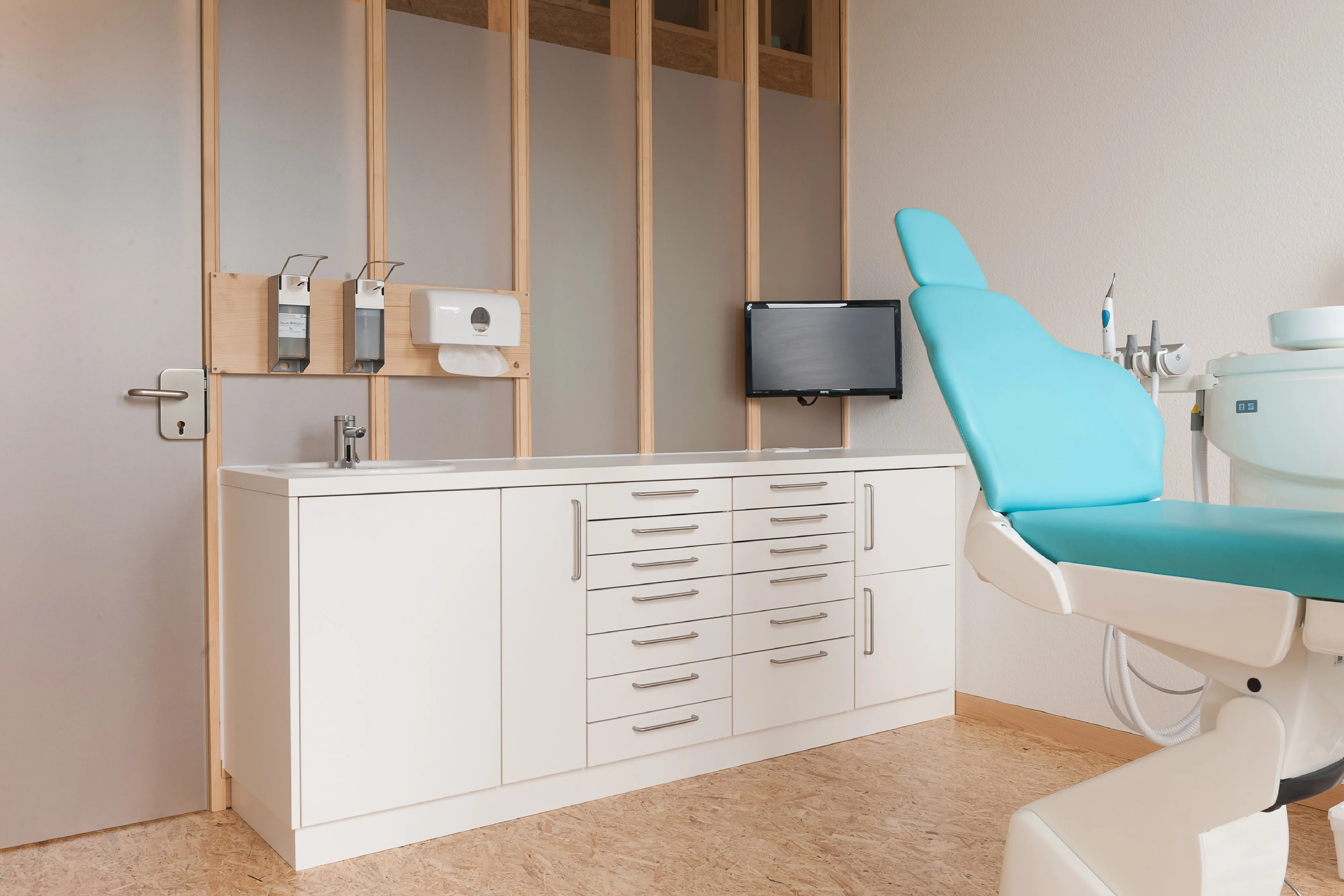 Behandlungsraum für Kinder mit einer maßgefertigten, weißen Unterschrankzeile vor einer hellen Raumteilerwand aus Holz und Mattglas.
