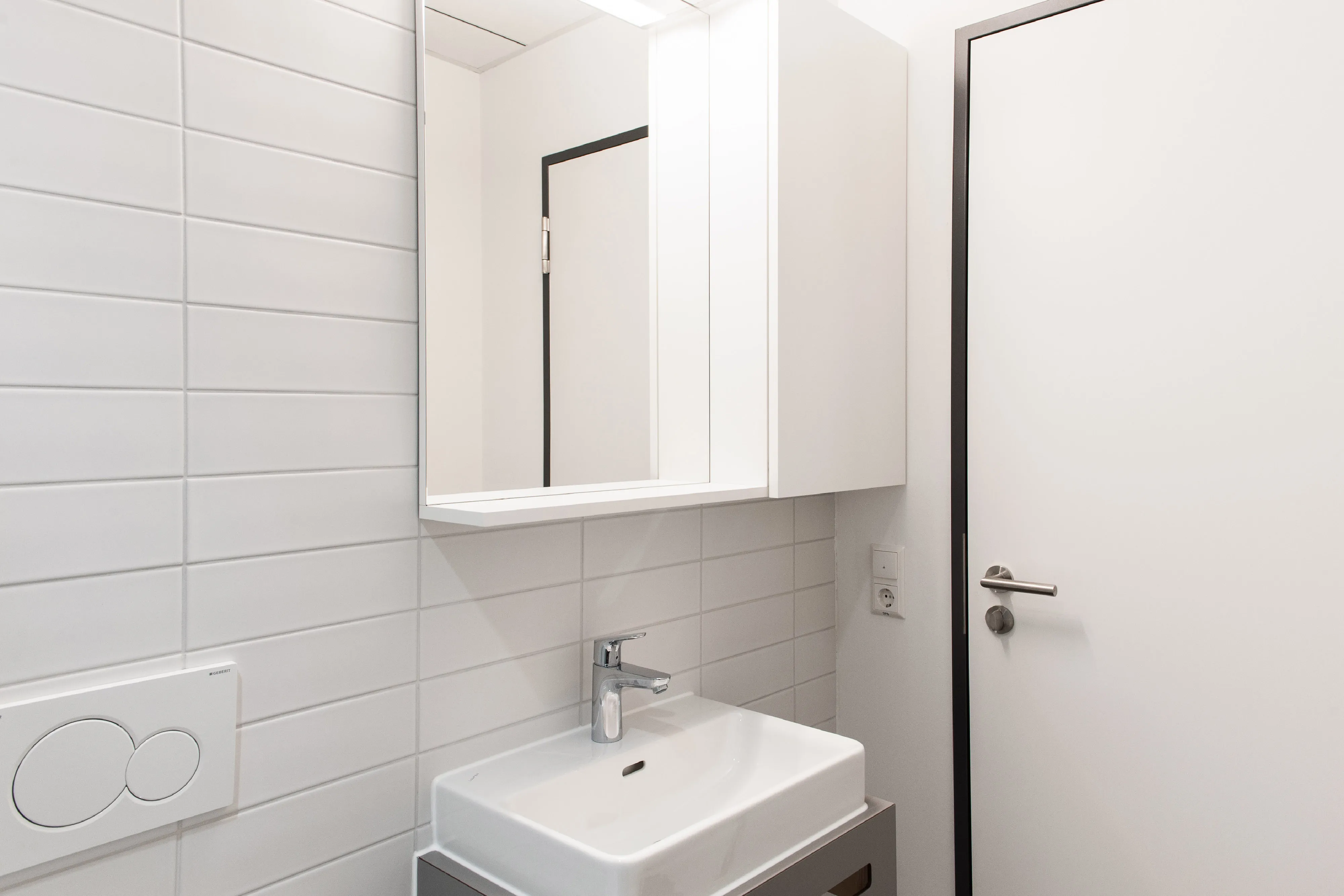 Hell gefliestes Kunden-WC mit modernem Waschplatz, grauem Waschtisch-Unterbau, Spiegelschrank und weißer Tür.