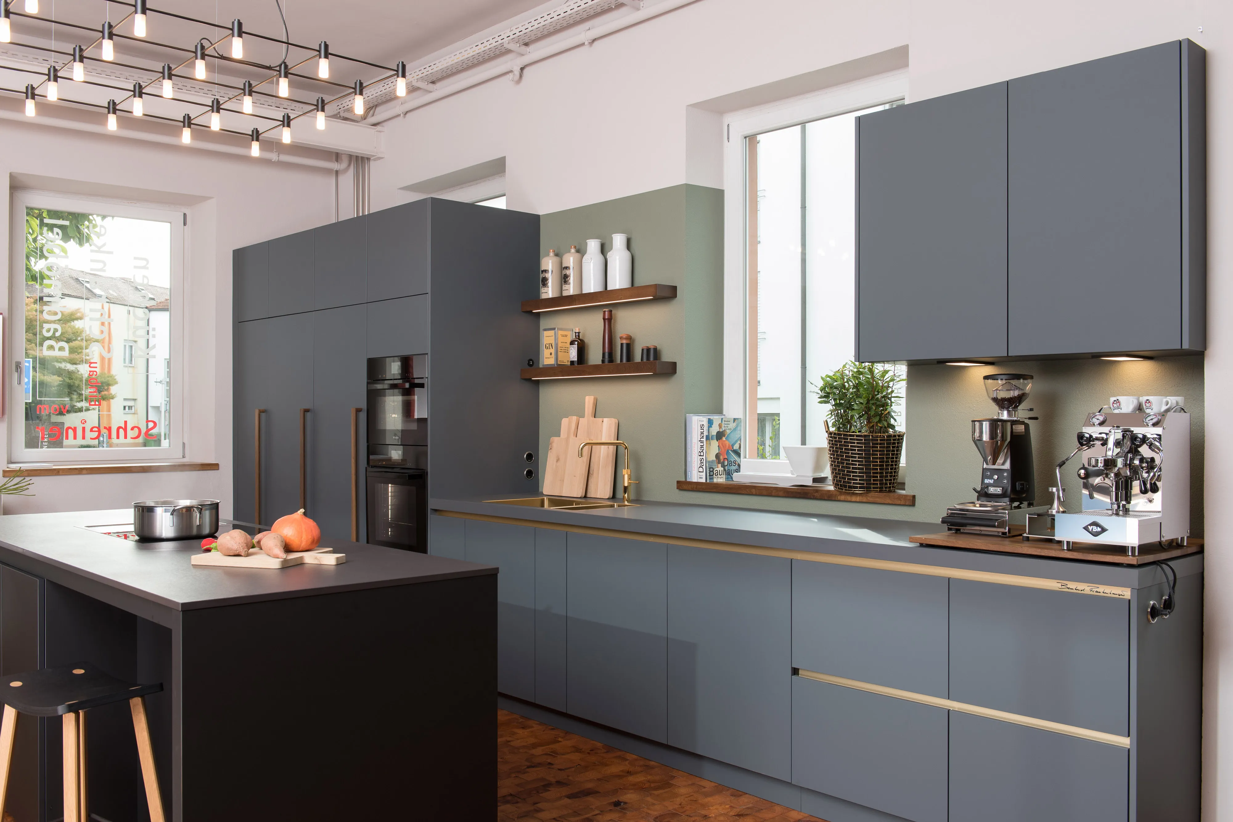 Ausstellungsküche im Industrial-Look mit matten dunkelgrauen Fronten, Arbeitsplatten, Holzregalen und einer Profi-Espressomaschine.