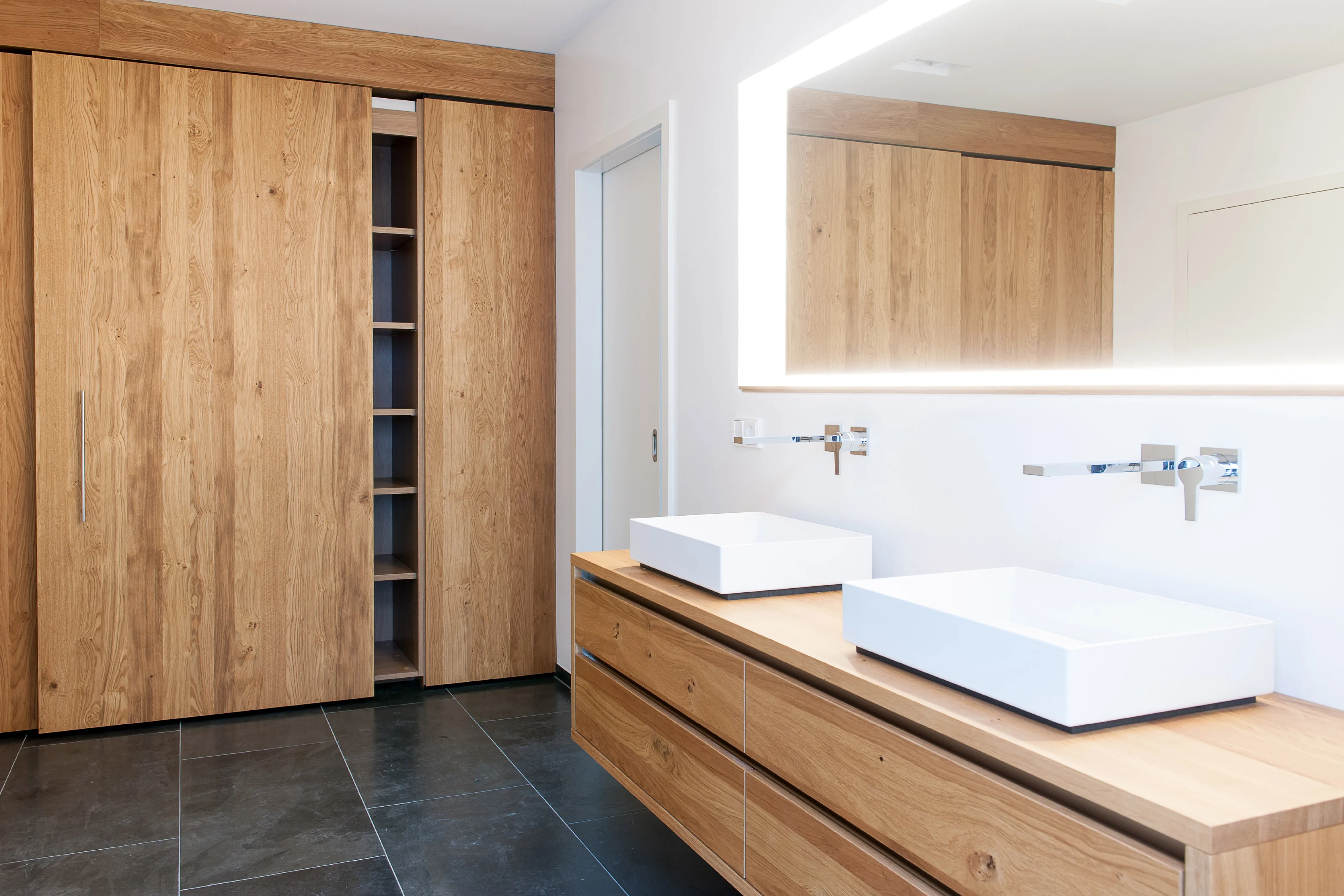 Helles Badezimmer mit maßgefertigtem Echtholz-Unterschrank, zwei eckigen Aufsatzbecken, beleuchtetem Spiegel und passendem Schiebetürschrank.