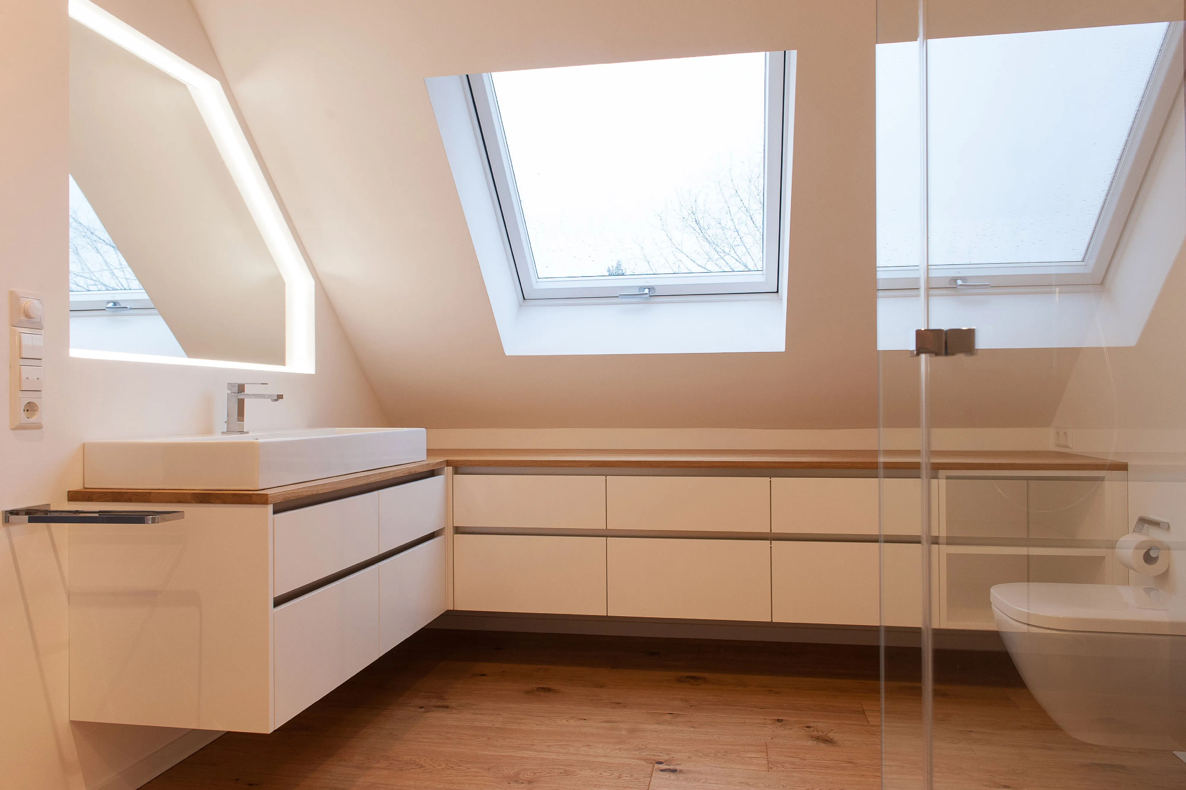 Dachgeschoss-Badezimmer mit weißem Waschtisch, Eichenholz-Abdeckung und maßeingepasstem Spiegelschrank unter der Dachschräge.