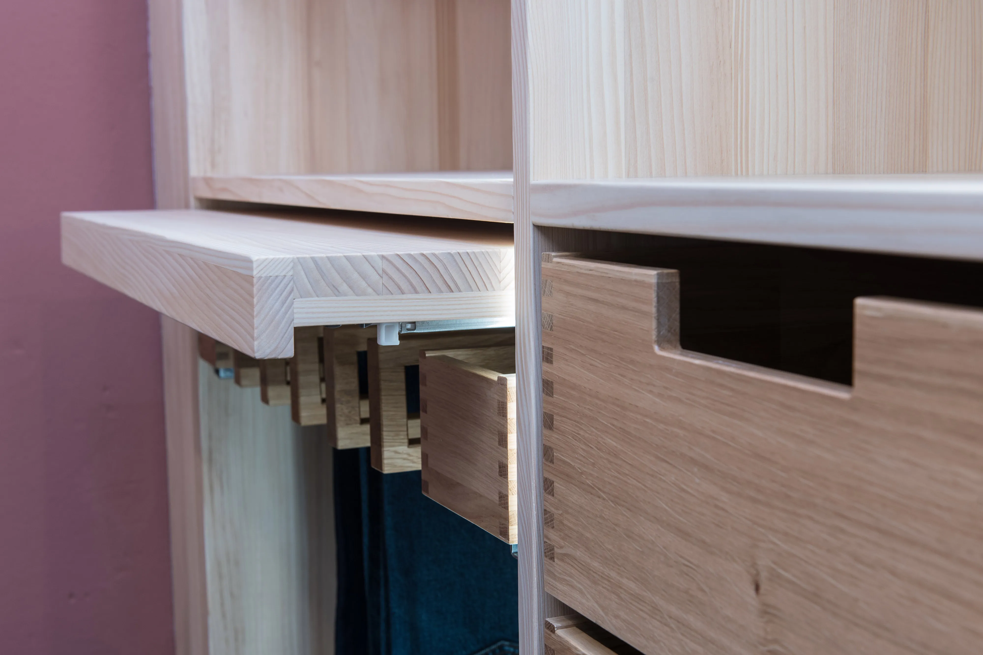 Detailansicht eines maßgefertigten Holzregalsystems mit ausziehbarer Hosenhalterung, gezinkter Holzschublade und LED-Beleuchtung.