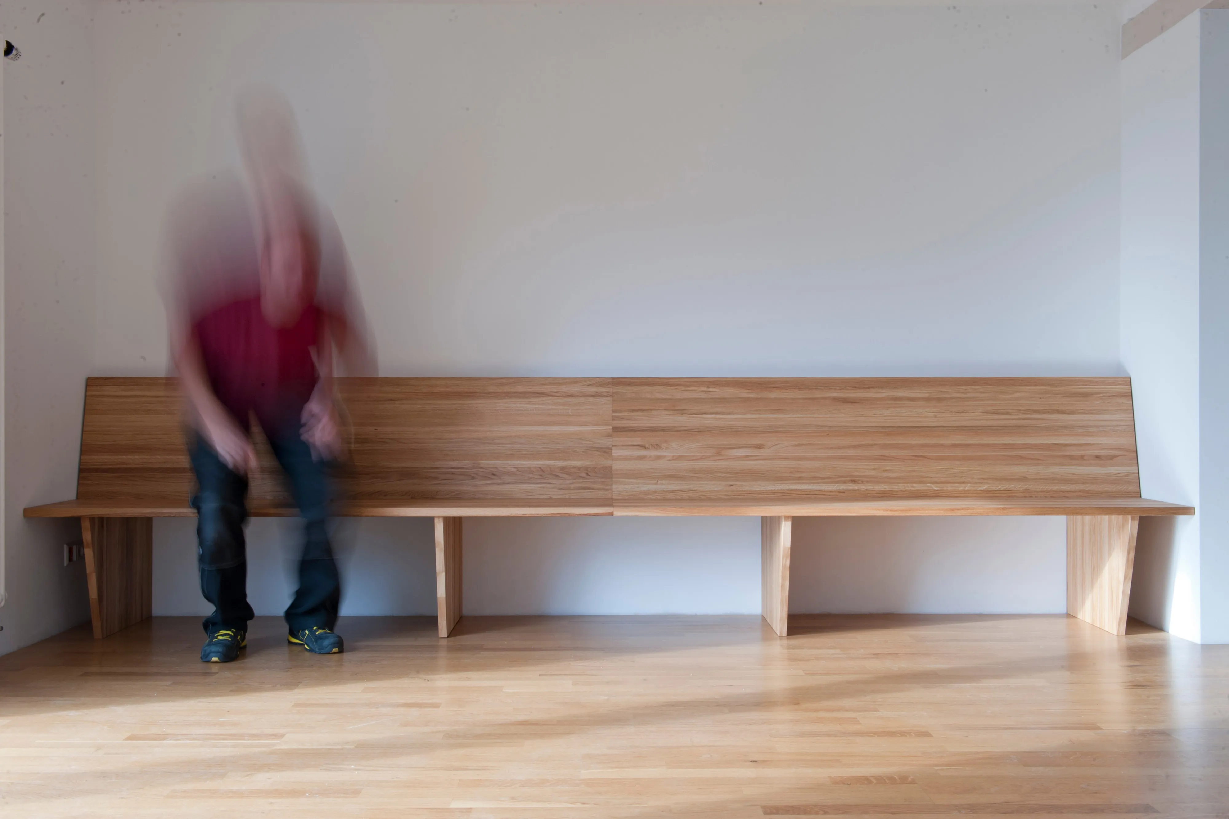 Aufgenommen im Bewegungsablauf: Mann setzt sich auf eine lange Holzbank an einer weißen Wand.