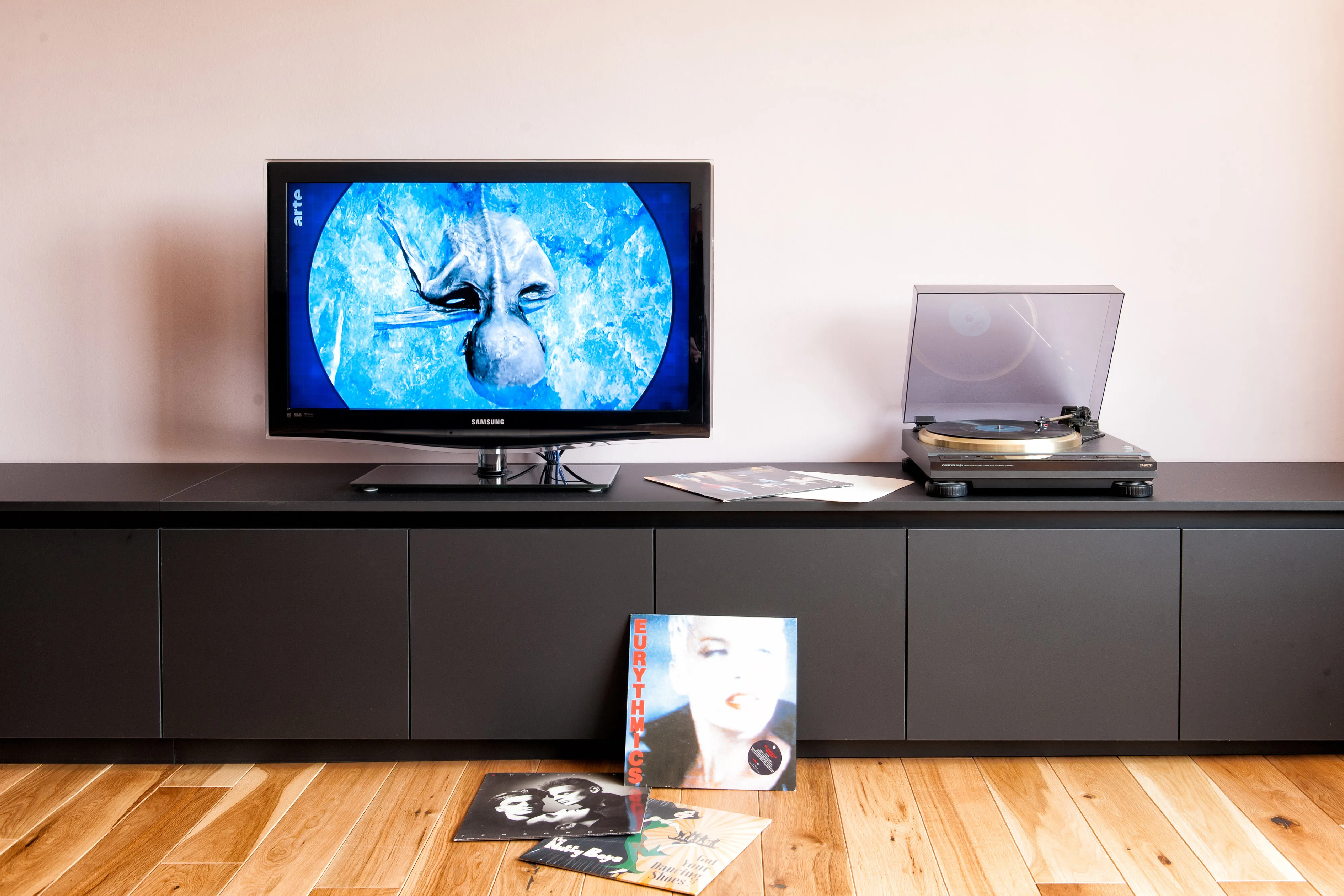 Niedriges, dunkelgraues Lowboard mit Fernseher, Plattenspieler und davor auf dem Holzfußboden platzierten Schallplatten.