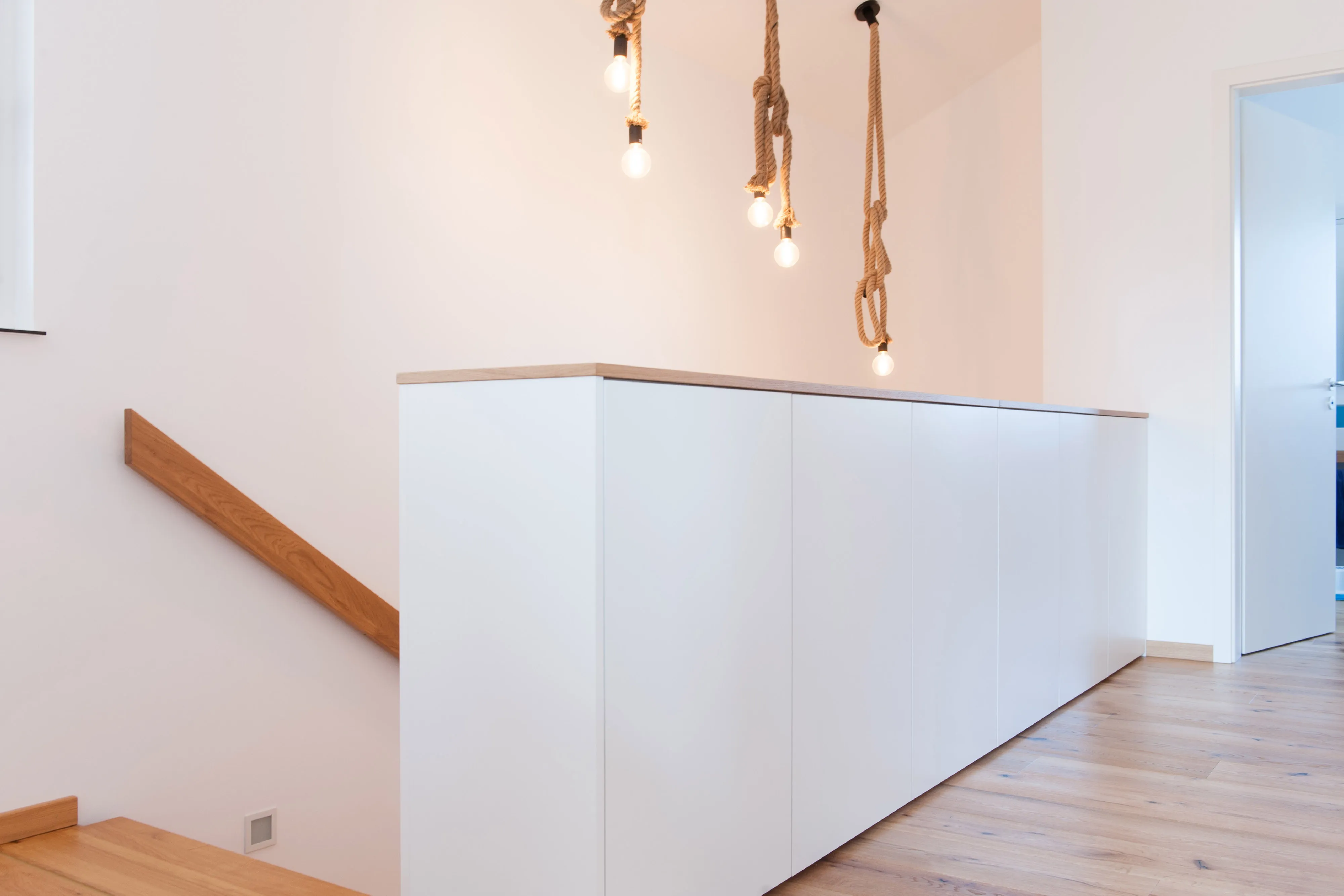 Weißes, griffloses Sideboard als Flur-Brüstung am Treppenaufgang mit hängenden Glühbirnen an Seilen.