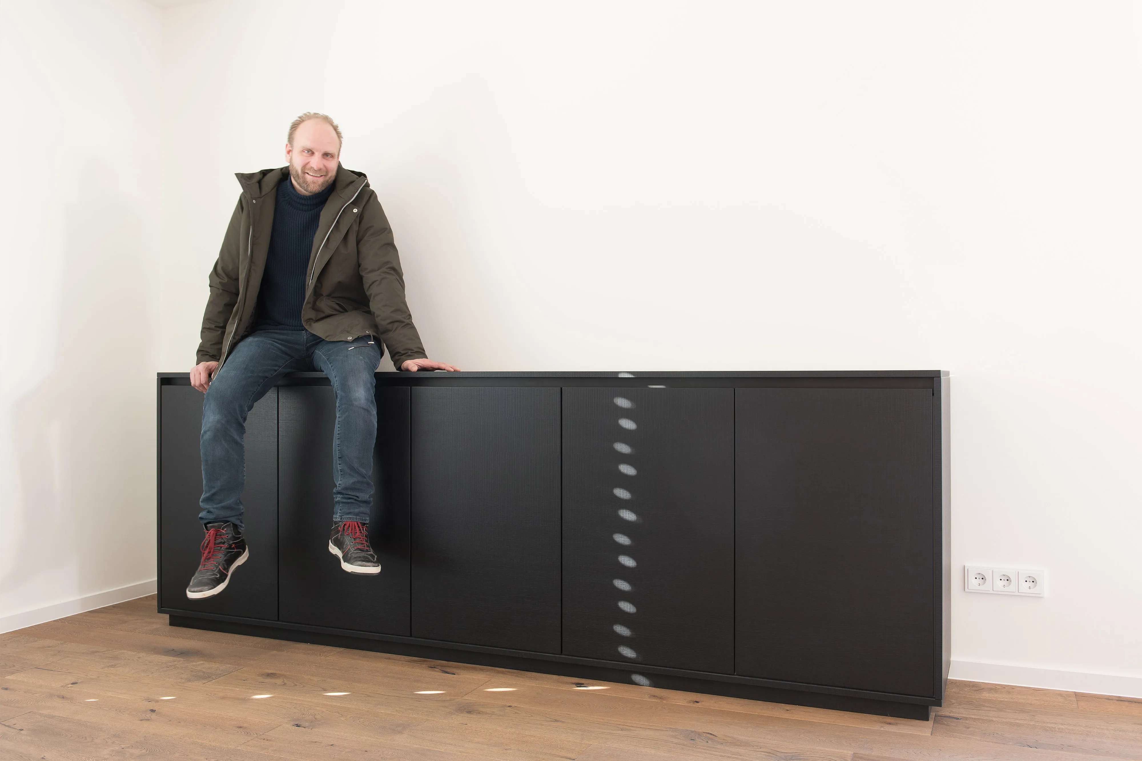 Bernhard Frankenhauser sitzt entspannt auf einem großen, dunkelbeizten Holz-Sideboard mit griffloser Front in einem leeren Raum.