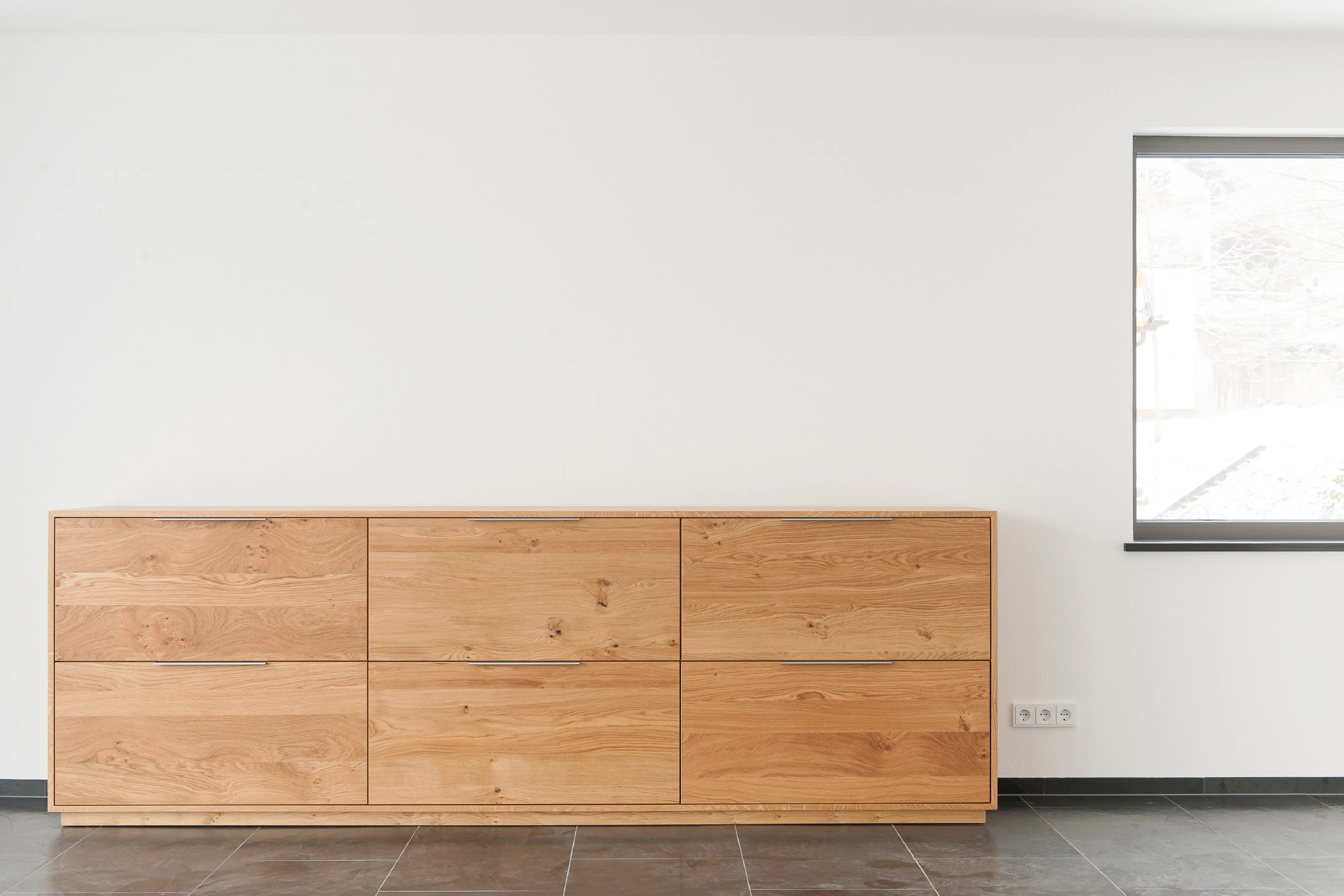 Minimalistisches Sideboard aus Echtholz mit sechs breiten Schubladen vor einer weißen Wand und dunklem Fliesenboden.