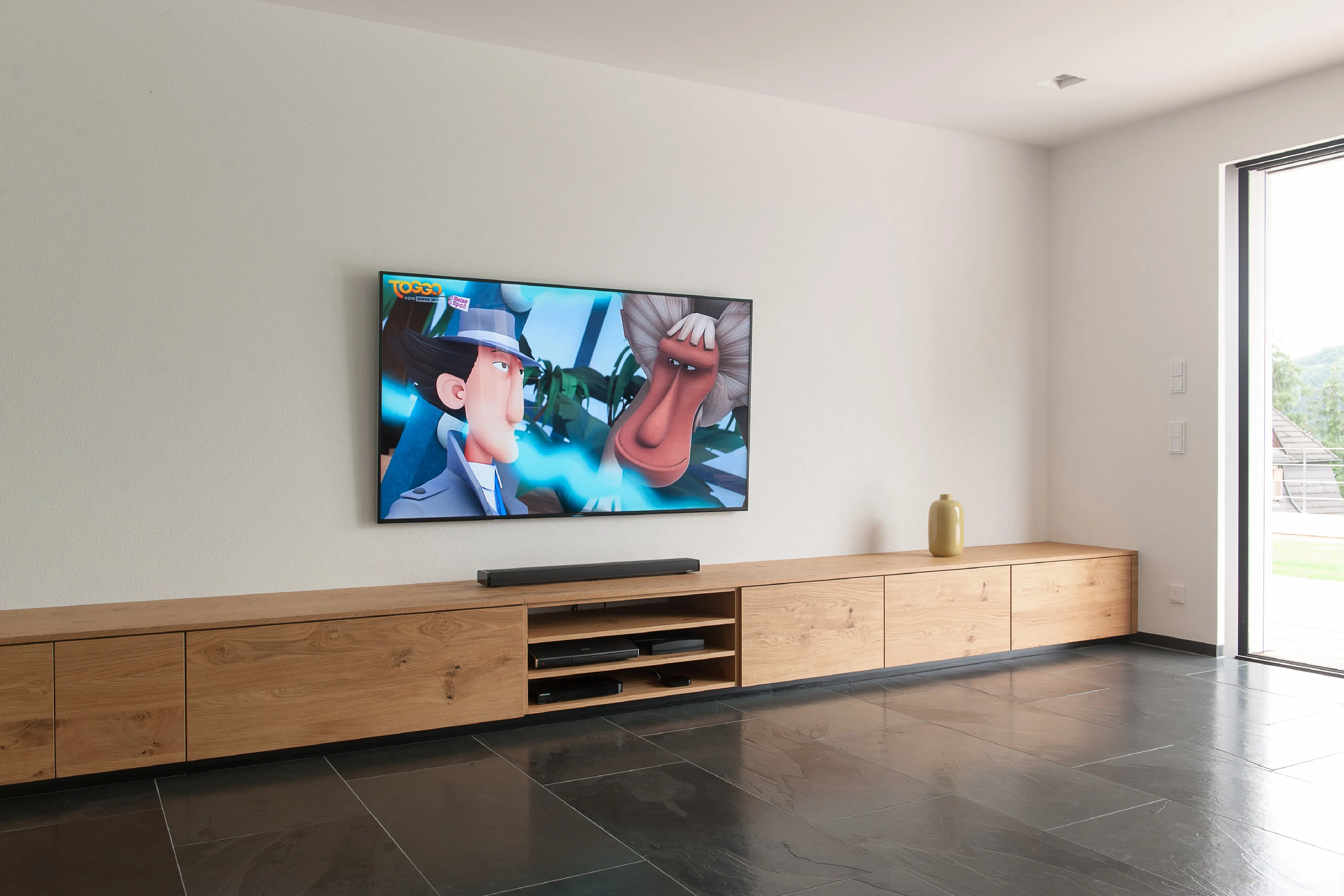 Sehr langes, niedriges TV-Lowboard aus Holz mit offenen Fächern für Hi-Fi-Geräte unter einem Wandfernseher.