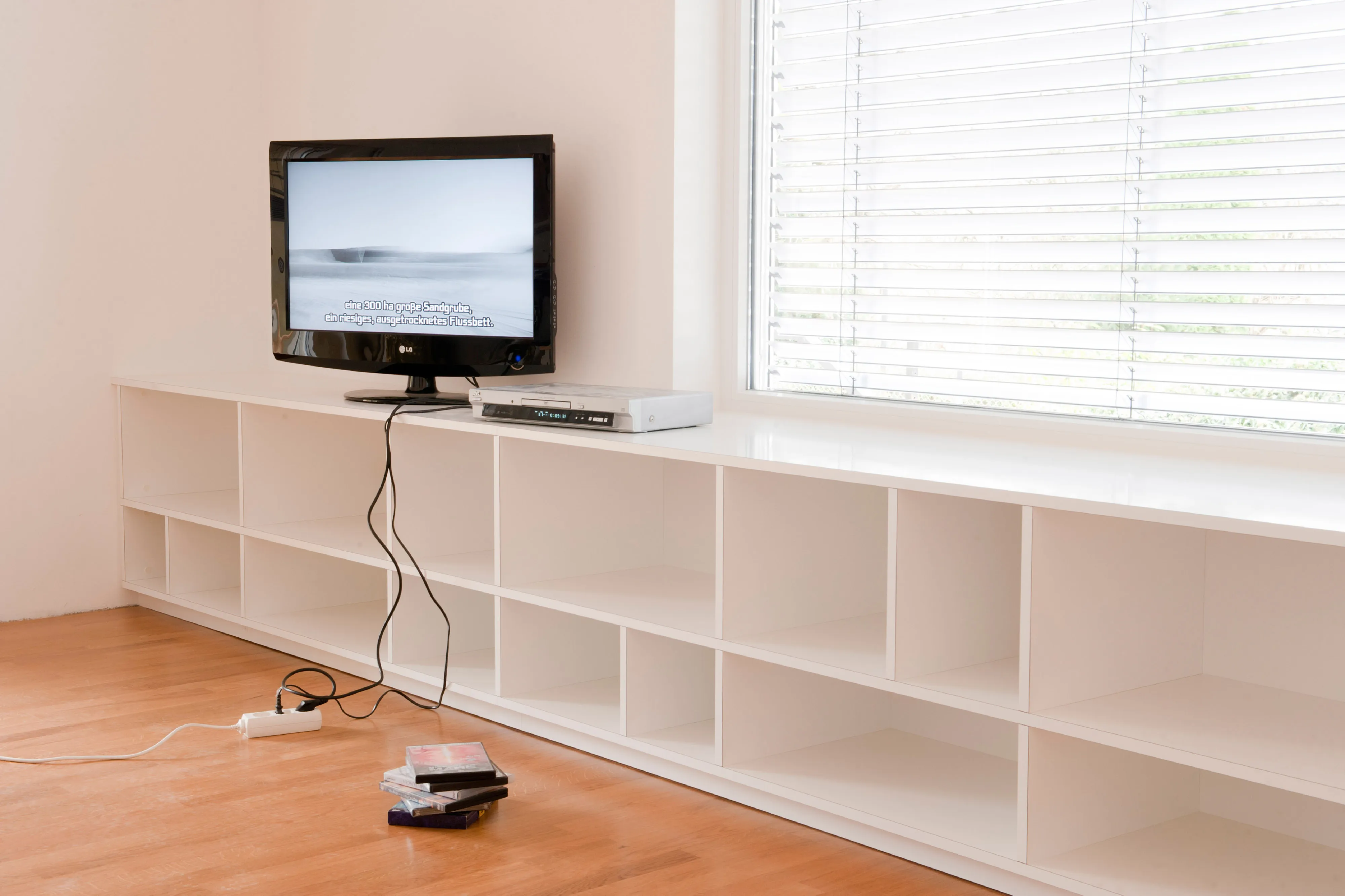 Flaches, weißes Sideboard mit offenen Fächern unter einem großen Fenster, genutzt als TV-Möbel mit Fernseher und DVD-Player.