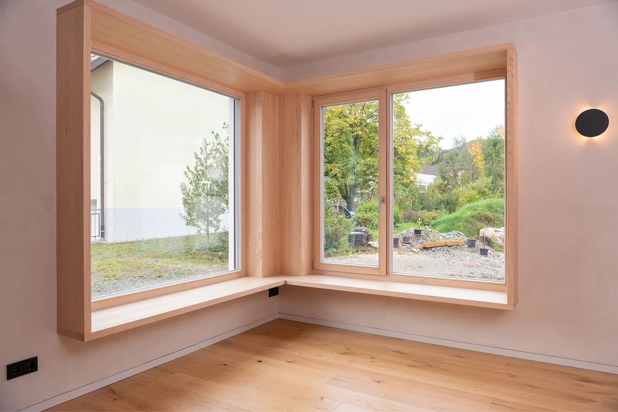 Maßgefertigtes Eck-Sitzfenster mit breitem Holzrahmen und tiefer Sitzfläche aus hellem Holz mit Blick ins Grüne.