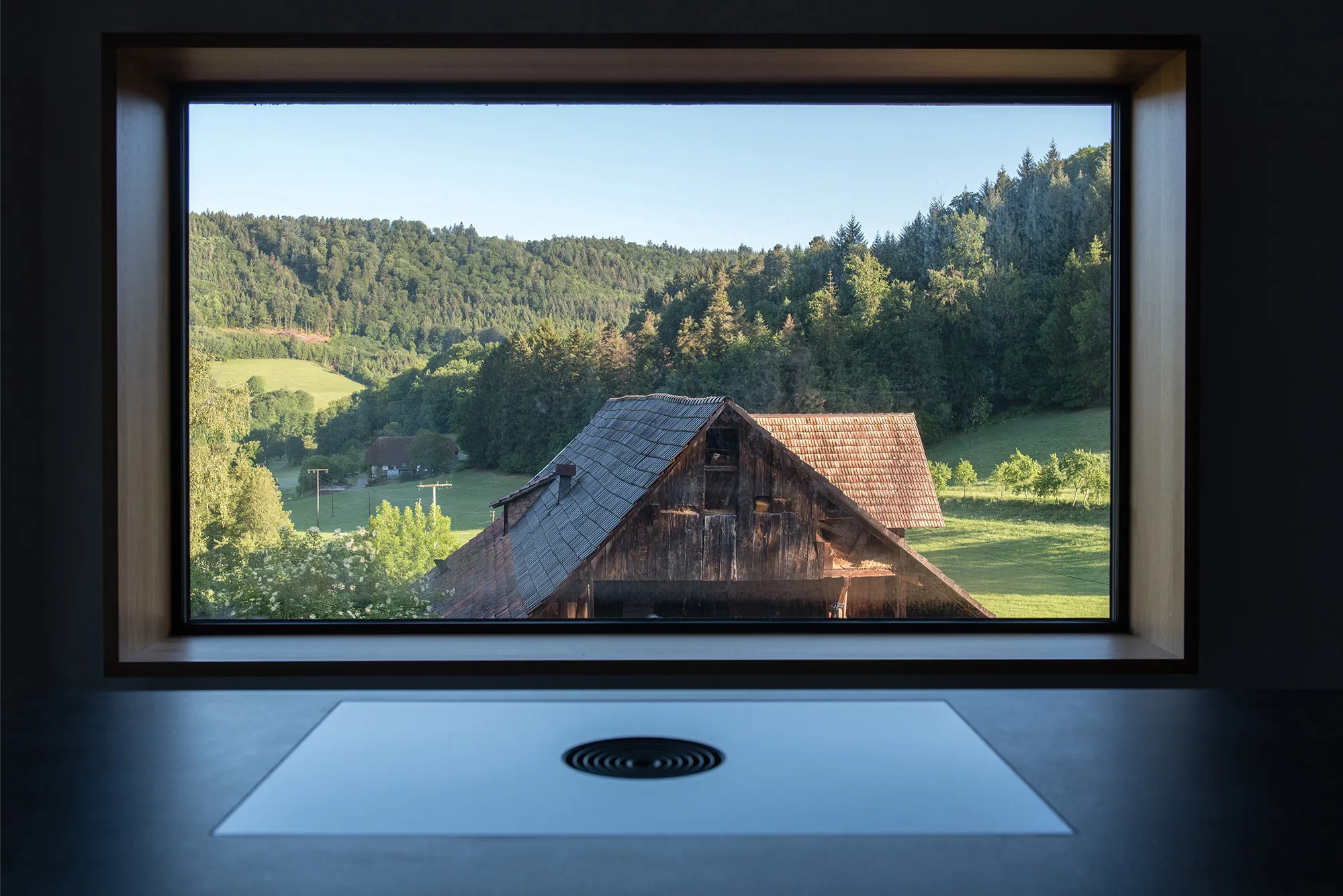 Panorama-Fenster mit tiefem Holzrahmen als Sitzbank mit freiem Blick auf eine grüne Landschaft und ein altes Schwarzwaldhaus.