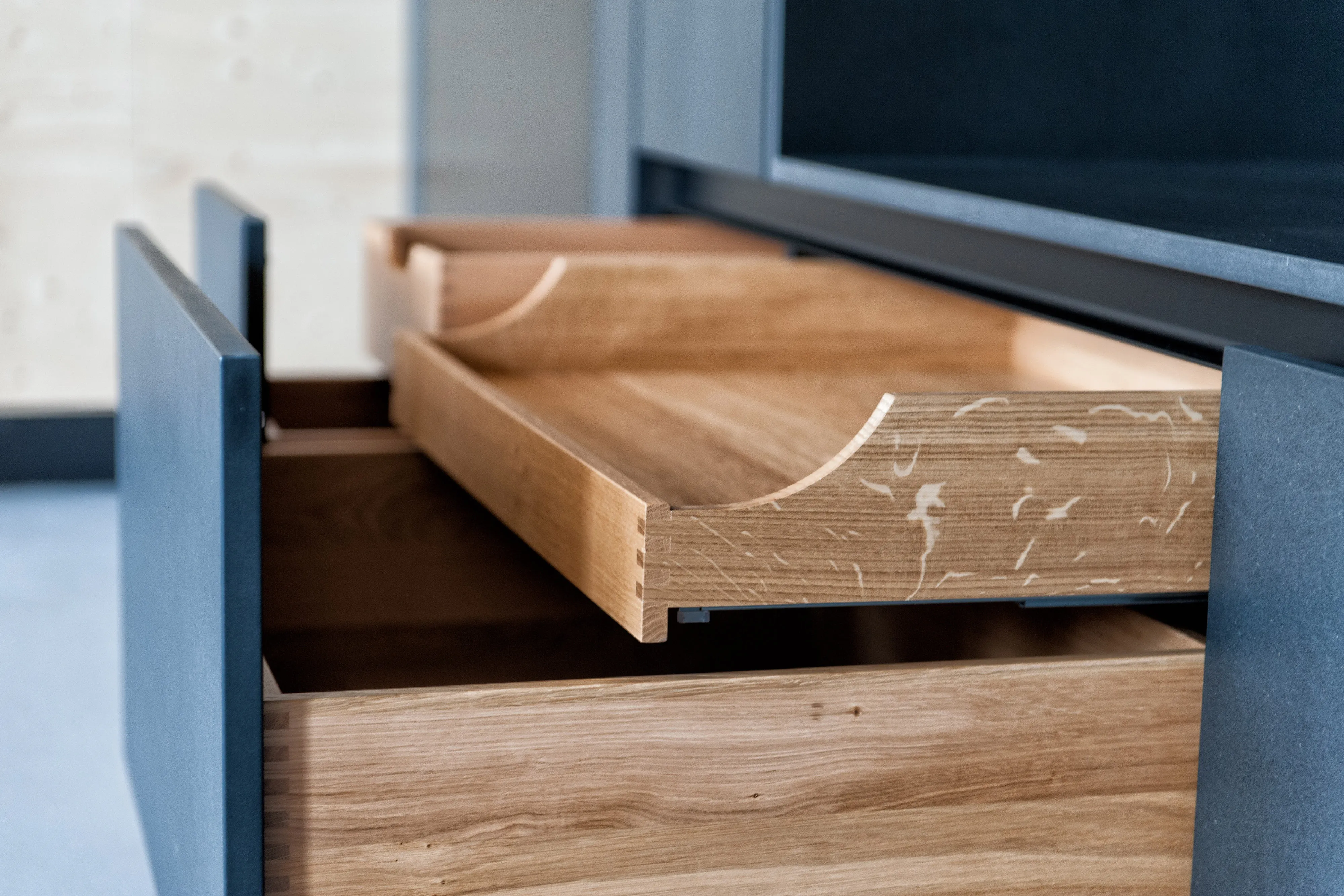 Detailansicht von geöffneten Massivholz-Schubladen mit geschwungenen Seitenwänden hinter dunkelgrauen Möbelfronten.