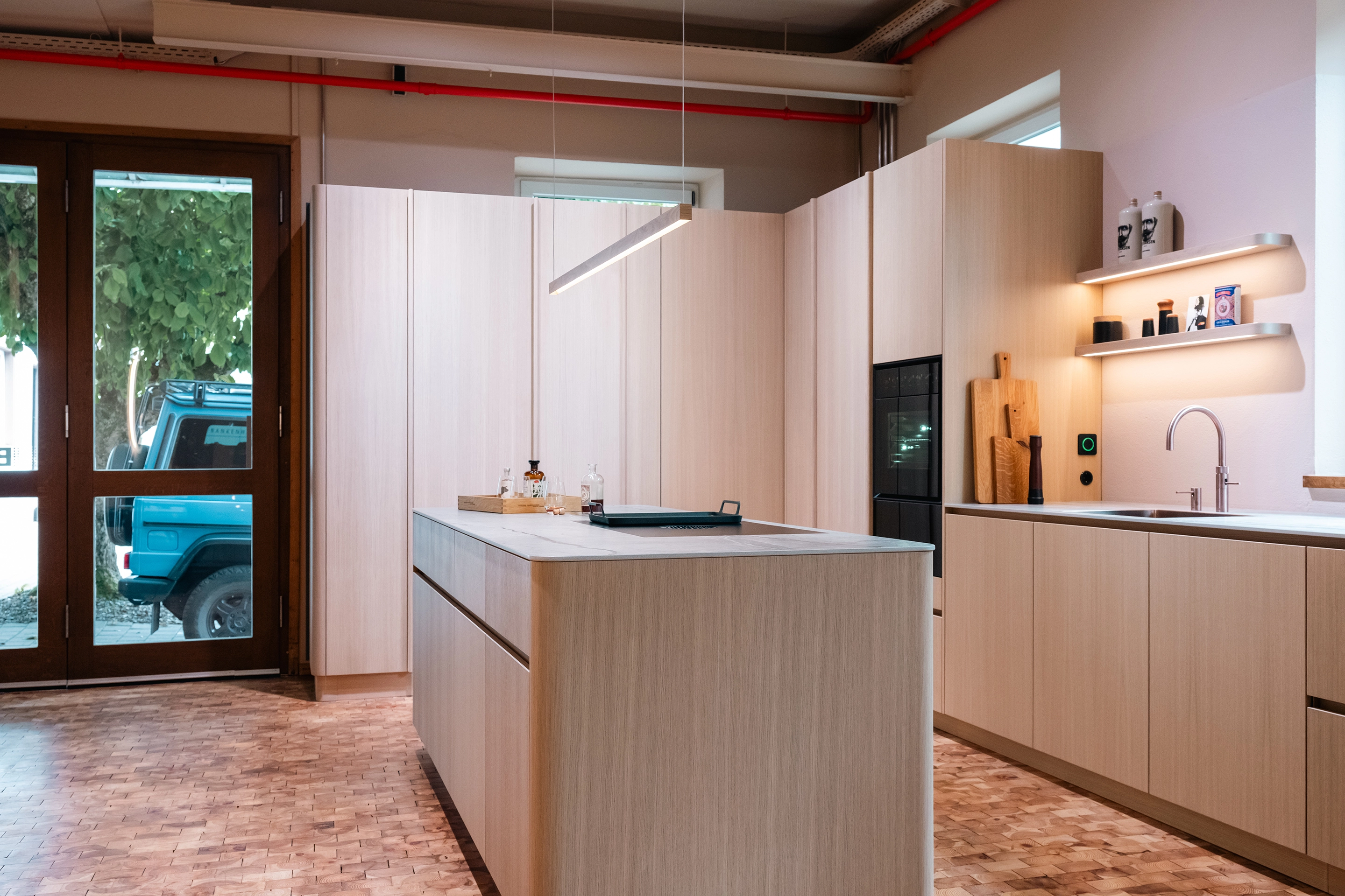 Modernes Küchenstudio aus hellem Holz mit Dingerstone Arbeitsplatte, BORA Backofen, Quooker Armatur und Hera Lichtleisten.