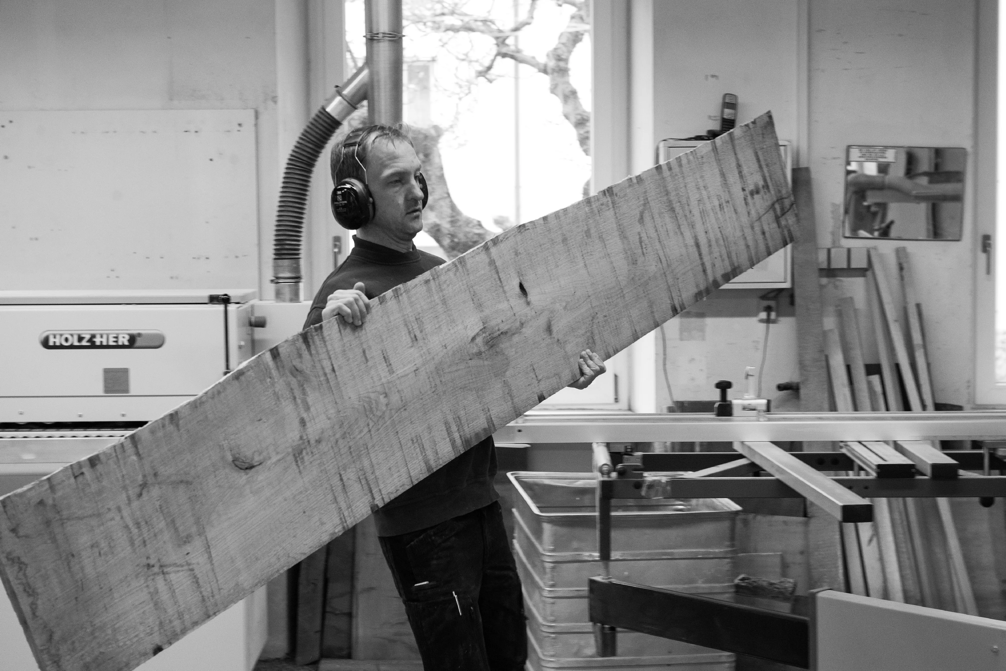 Schwarz-Weiß-Fotografie eines Schreiners mit Gehörschutz, der ein großes, massives Holzbrett durch die Maschinenhalle trägt.