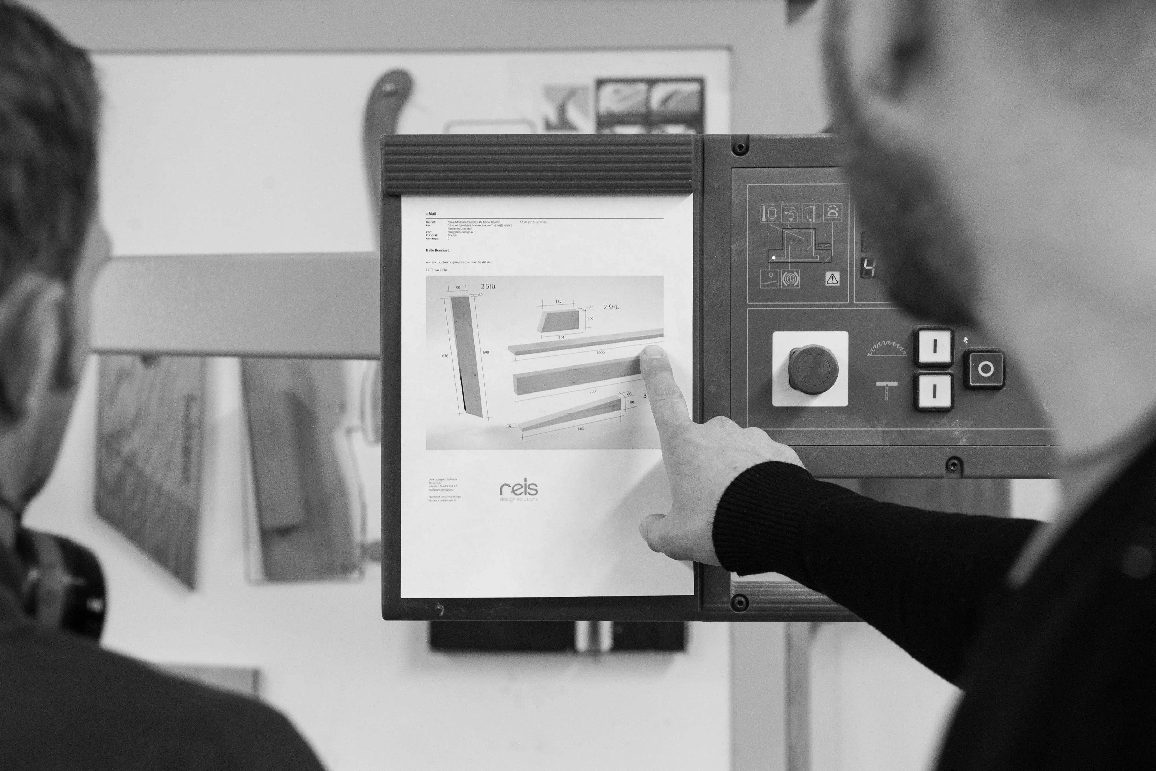 Schwarz-Weiß-Ansicht zwei Schreiner, die auf ein Display einer Maschine schauen und eine Technische Zeichnung besprechen.