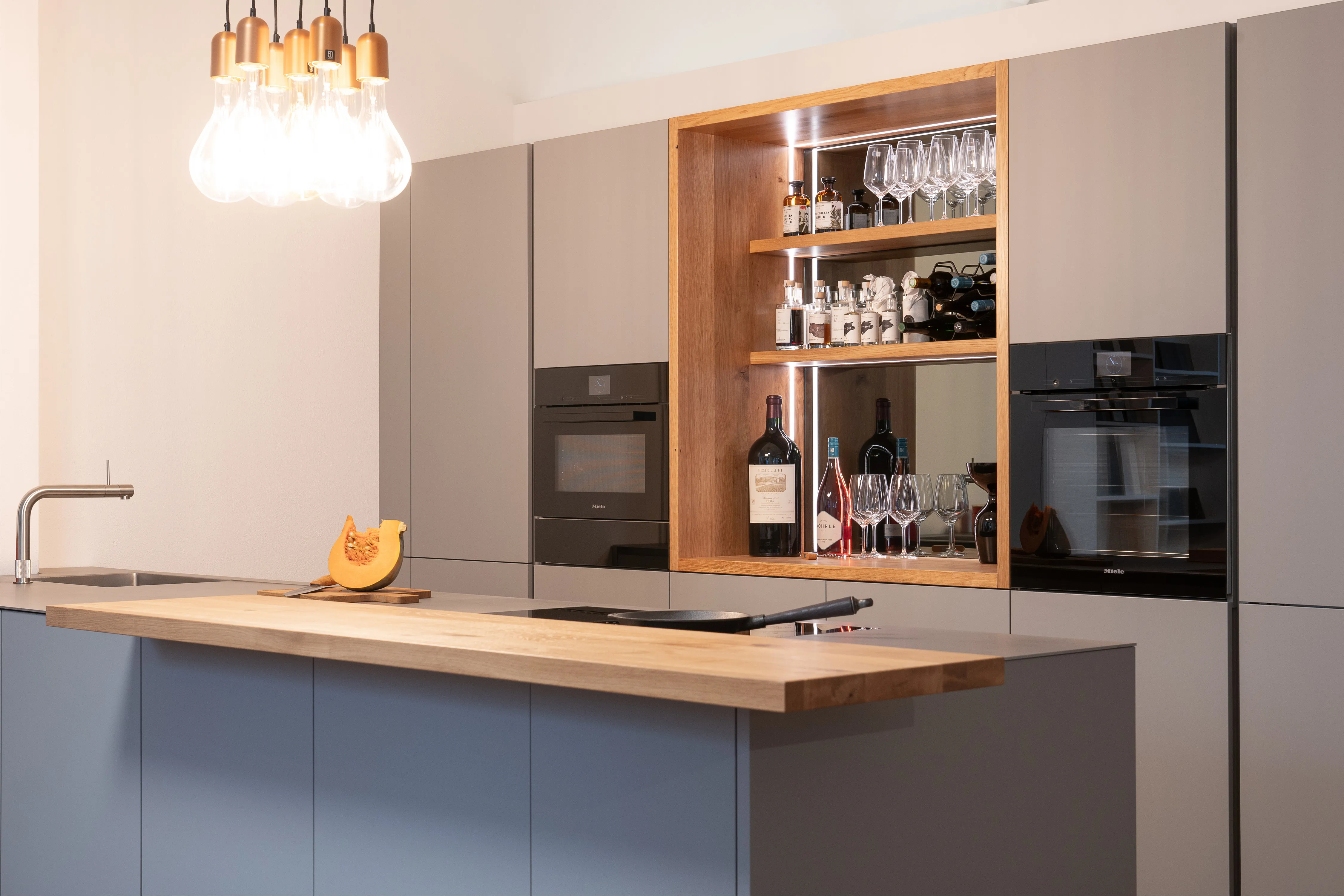 Graue Kücheninsel mit Eichenholz-Theke, Kochfeld und Hochschrankwand mit integriertem Holz-Barregal