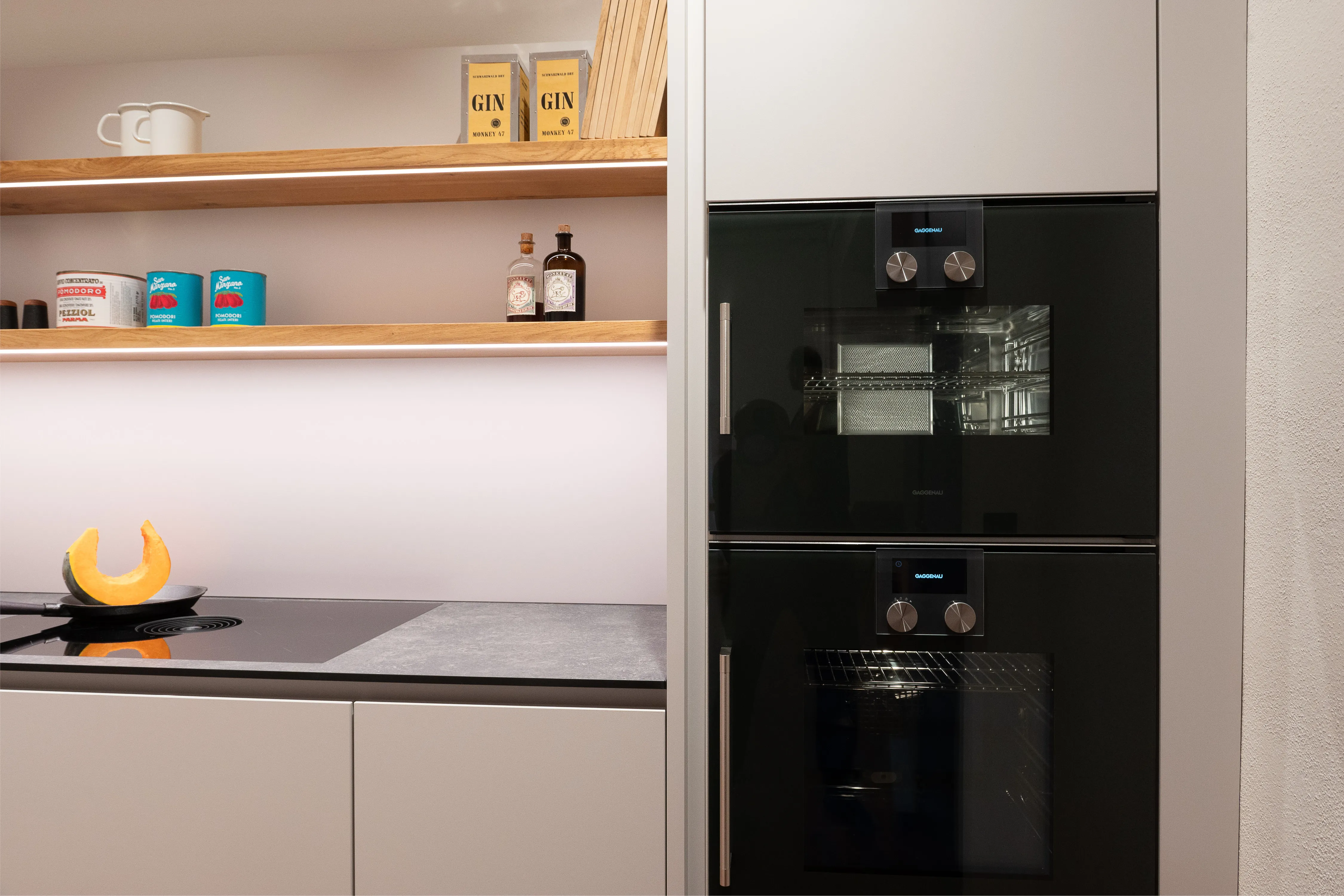 Detailansicht zweier übereinander eingebauter schwarzer Gaggenau-Backöfen neben beleuchteten Holzregalen